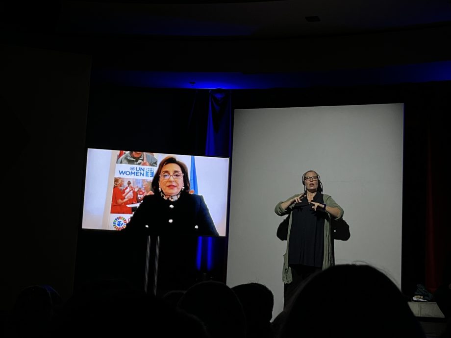 On the left of the picture is a screen on which a woman's video message is being transmitted. To the right, a Sign Language interpreter stands in the event room and translates to the audience