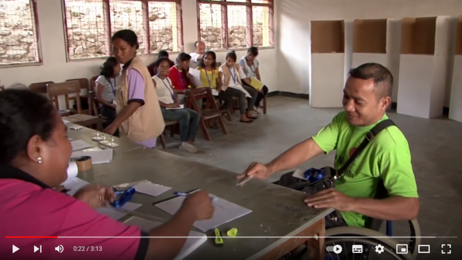 A man in a wheelchair approaches the registration table at an election office. This video will open in YouTube.