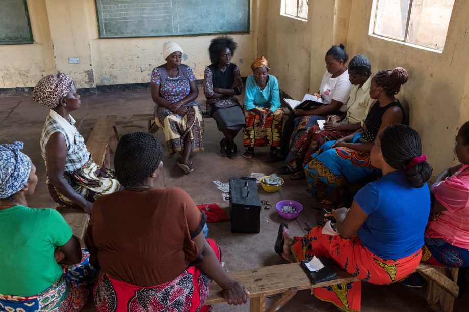 A group of adults sits in a circle on wooden benches in a classroom. In the middle are various things, e.g. notepads and a large cash box.