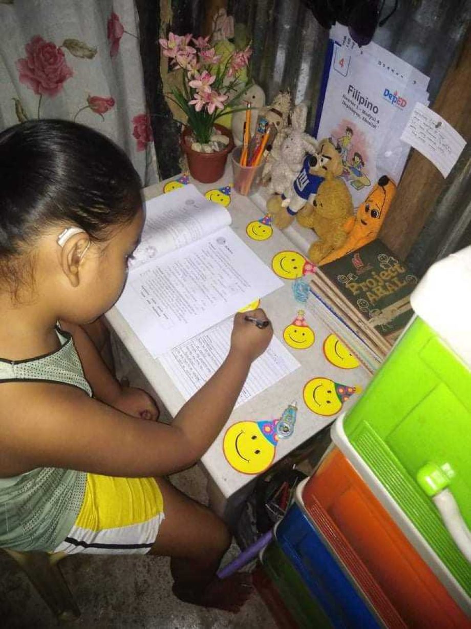 Franco sits barefoot at his small desk, which is decorated with many yellow smiley stickers. He has an exercise book and a sheet of paper in front of him. A row of cuddly animals sits on his desk and watches Franco write.