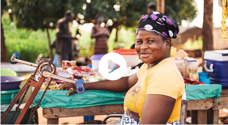 A woman is sitting on a tricycle. You can see her from the side and only up to under her chest. She turns her head to the camera and grins. In the background a market stall can be seen out of focus.  This video opens on YouTube.