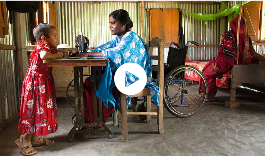 A woman sits at a sewing machine. Opposite her leans a little girl. A wheelchair can be seen in the background. This video opens on YouTube.